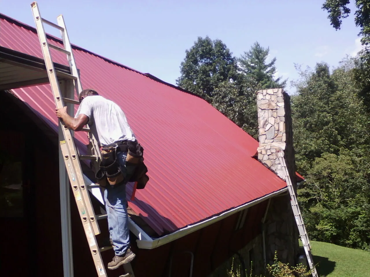 Roofer on a ladder beside a red metal roof and stone chimney — Soffit & Fascia Repair by Roofing Brothers in Southbridge