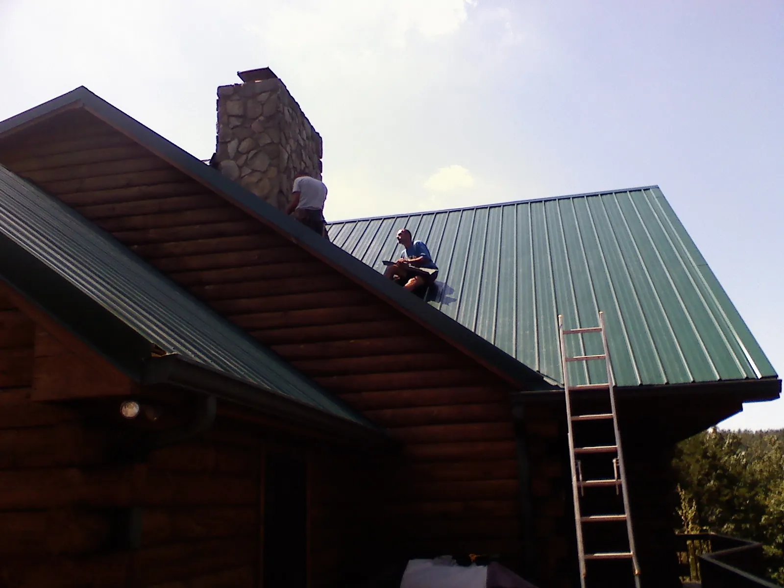 Roofers installing green metal panels beside a stone chimney on a log home in Southbridge